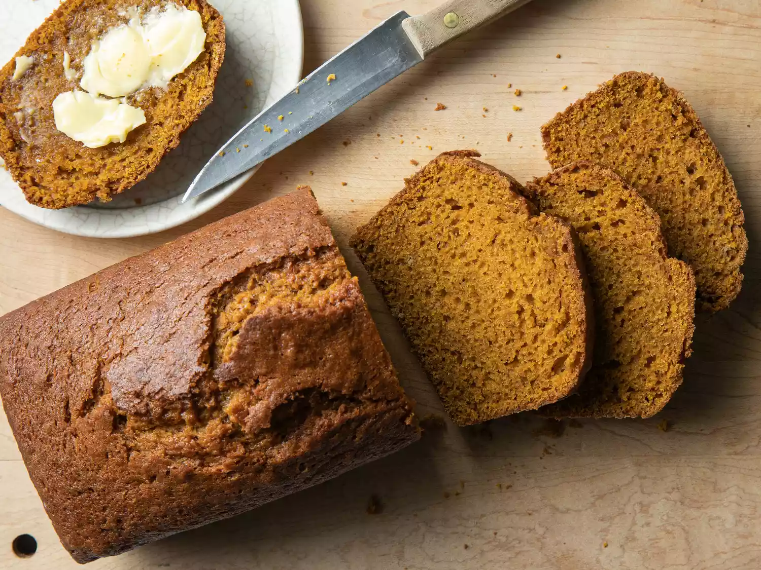 an overhead view of partially sliced pumpkin bread sitting on a wooden cutting board with a single slice buttered.