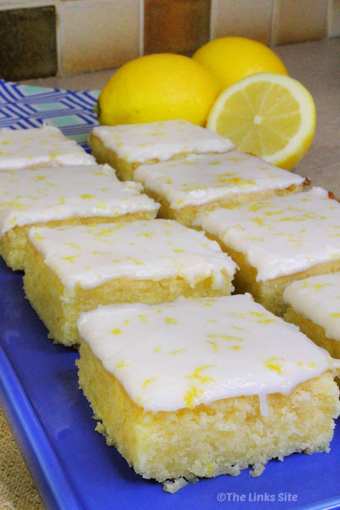 Eight iced lemon brownies, arranged in two rows of four on a blue plate. A blue and white patterned tea towel, two whole lemons, and a half of a lemon can be seen in the background.